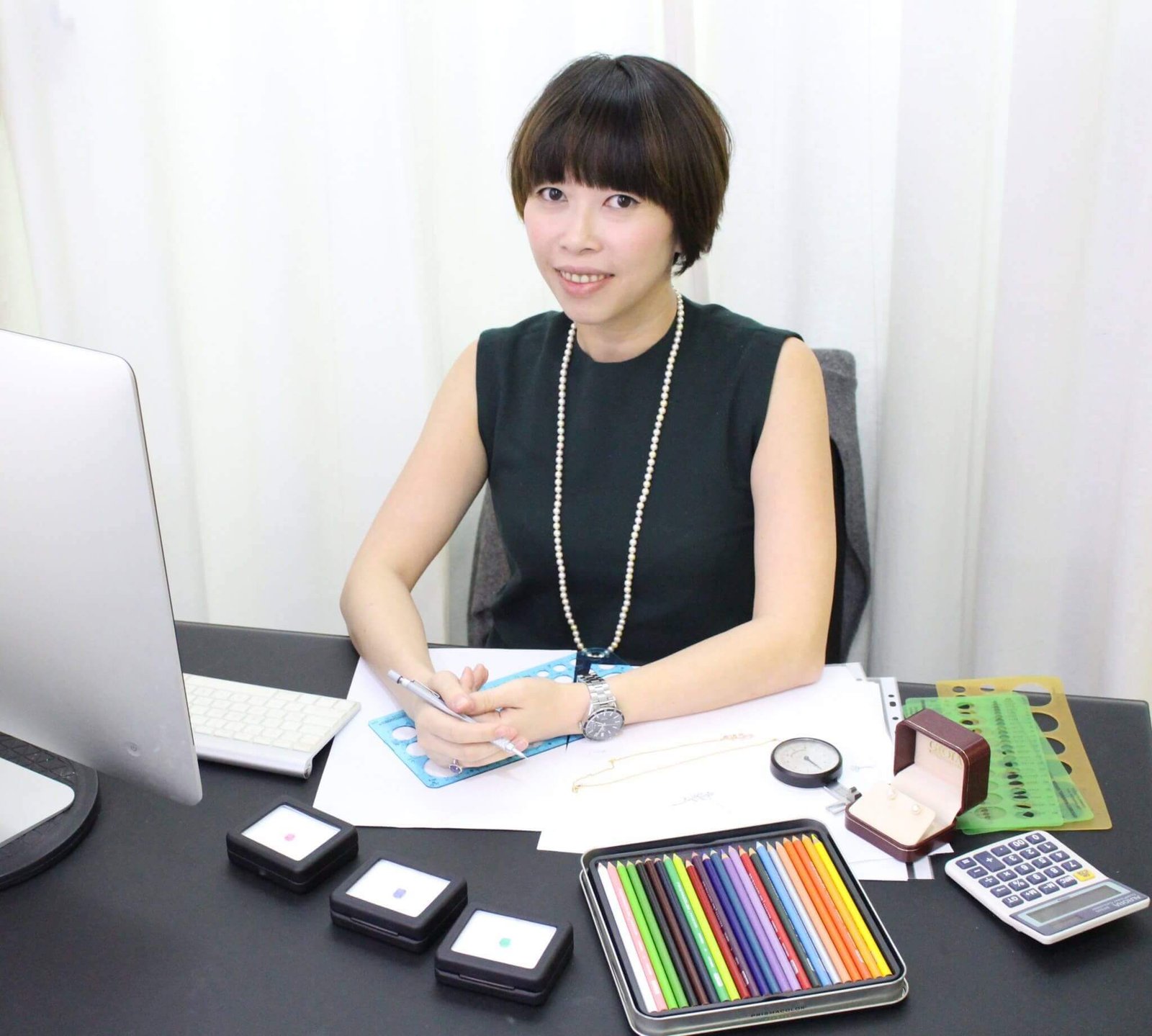 Cheryl Woon sketching a jewellery design at her desk, with gemstone comparison boxes and coloured pencils, GIOIA Fine Jewellery