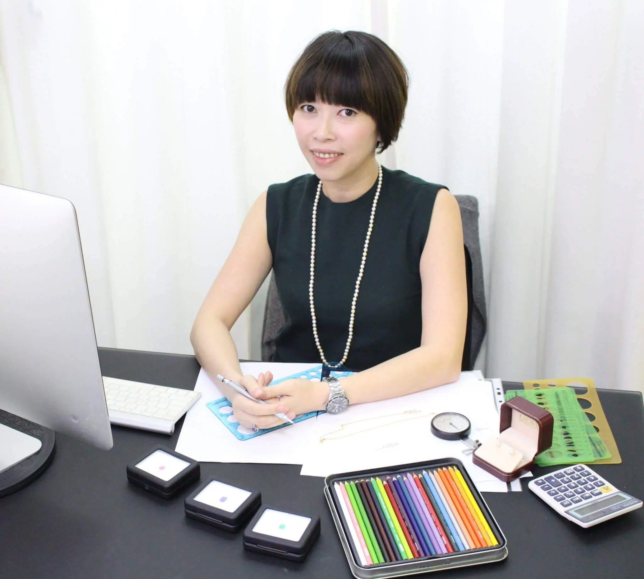 Cheryl Woon sketching a jewellery design at her desk, with gemstone comparison boxes and coloured pencils, GIOIA Fine Jewellery
